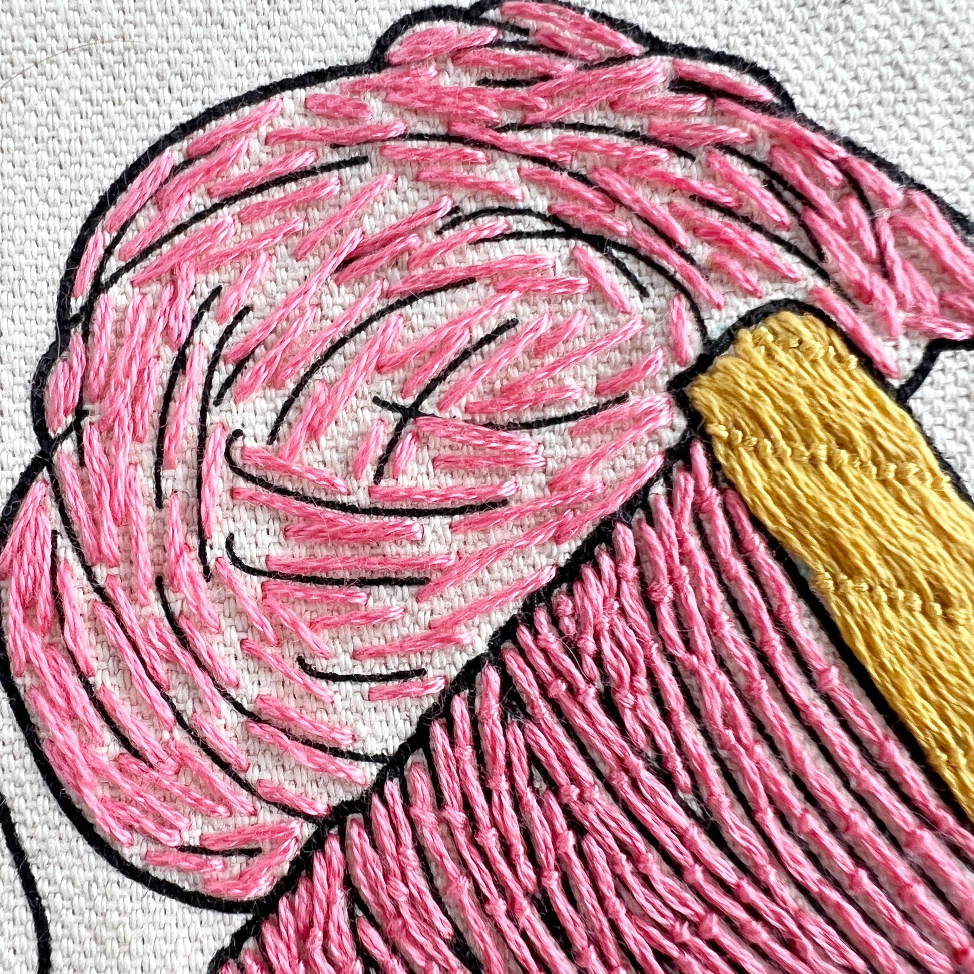 Close up of drop spindle and roving embroidered in pink on Rosanna Diggs Fiber Fluid tote.  Roving has long straight stitches.  Drop spindle has details in the woodwork  design embroidered in a yellow gold.