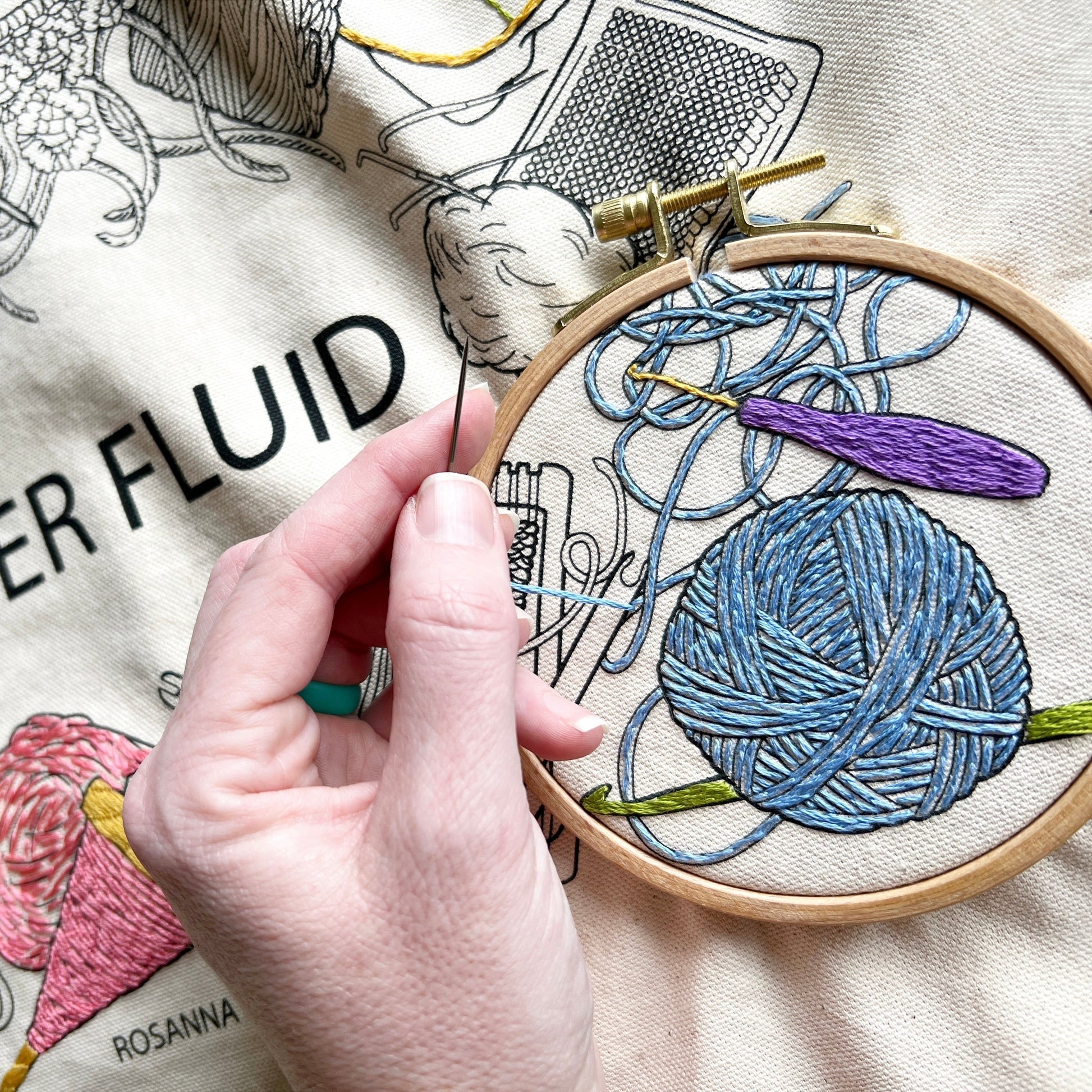 Close up of bag showing someone stitching a  ball of yarn that is unraveled surrounded by crochet hooks. Yarn ball is embroidered in blue.  Crochet hooks are green and purple.  Four inch hoop surrounds area being worked on.