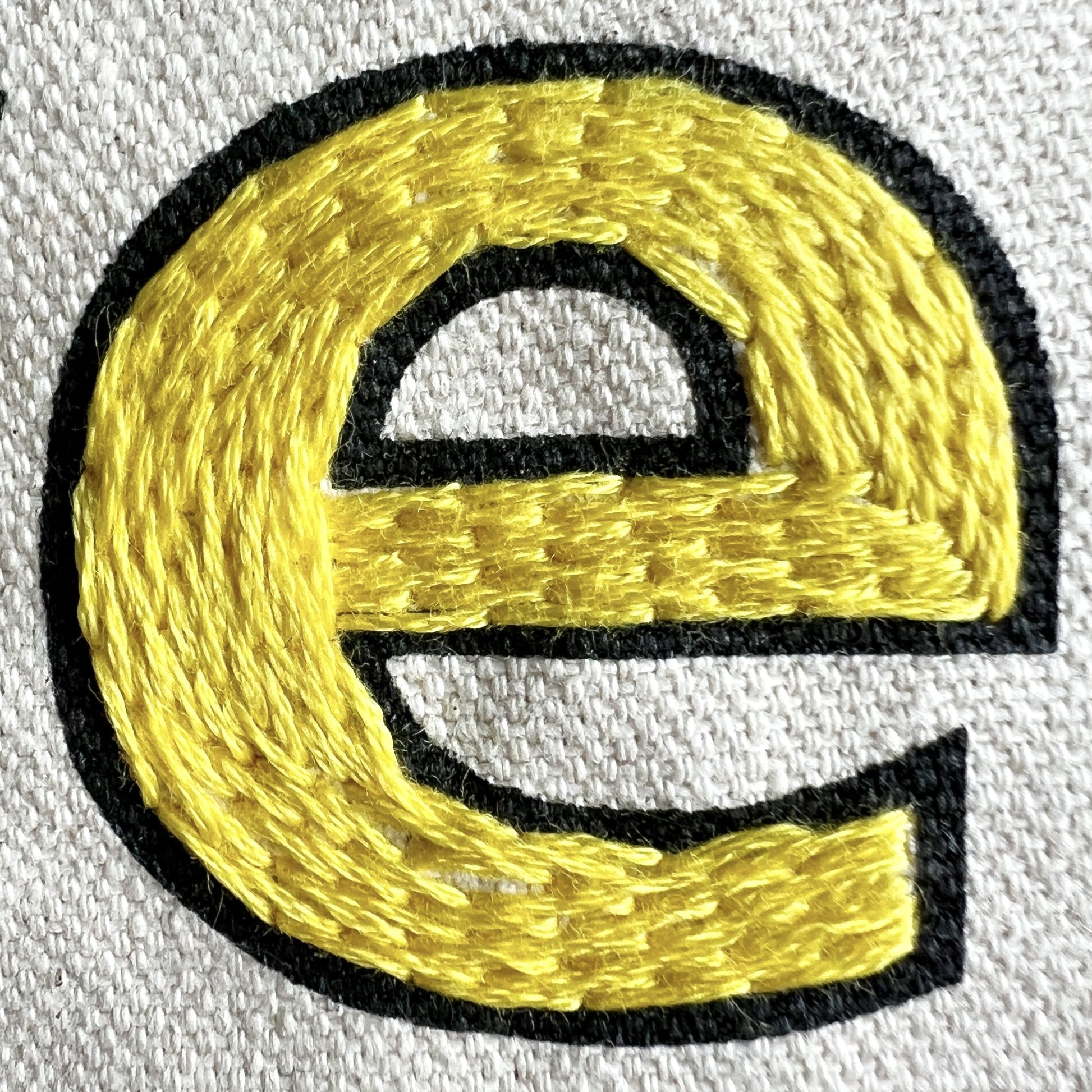 Close up stitch details for Love Your Neighbor Tote Embroidery Kit.  Black outlined letter on cream canvas is filled with bright yellow stitches resulting in an eye catching letter e.