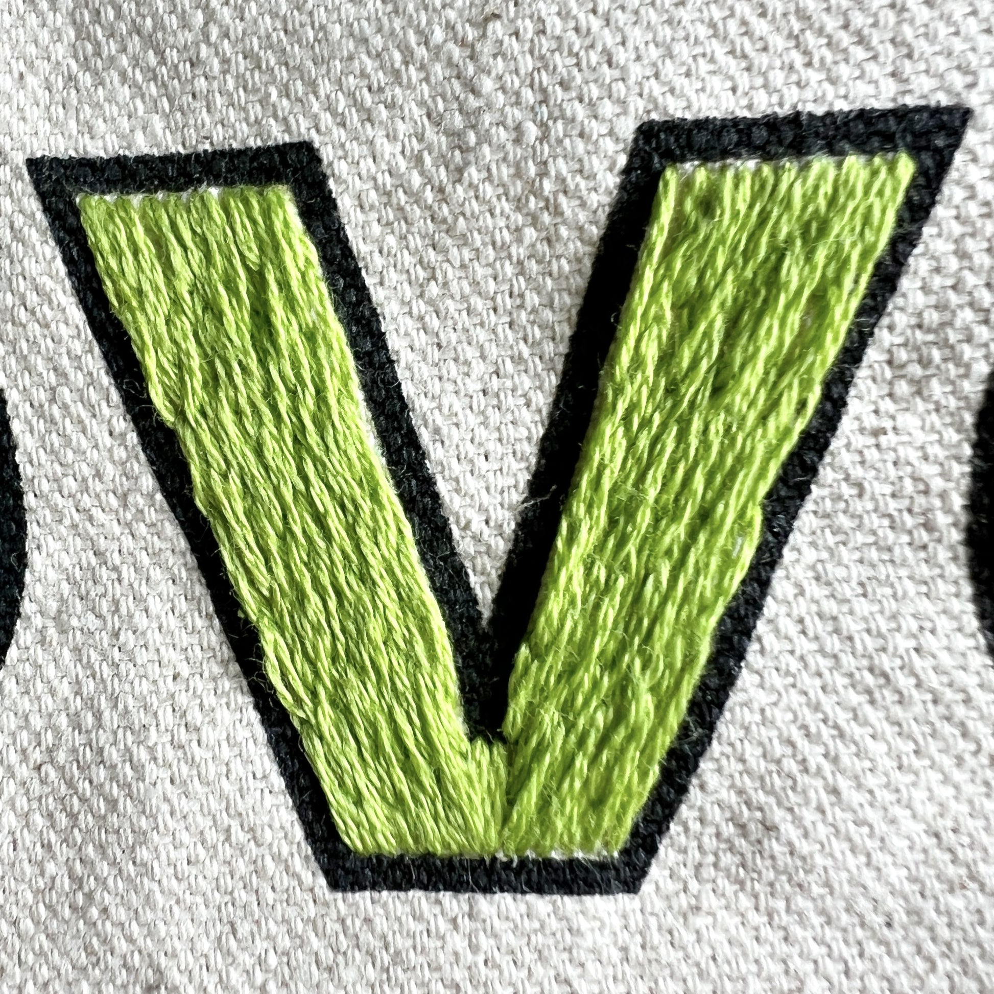 Close up stitch details for Love Your Neighbor tote bag embroidery kit. Letter V open block printed on cream canvas filled with green stitching for a solid letter.