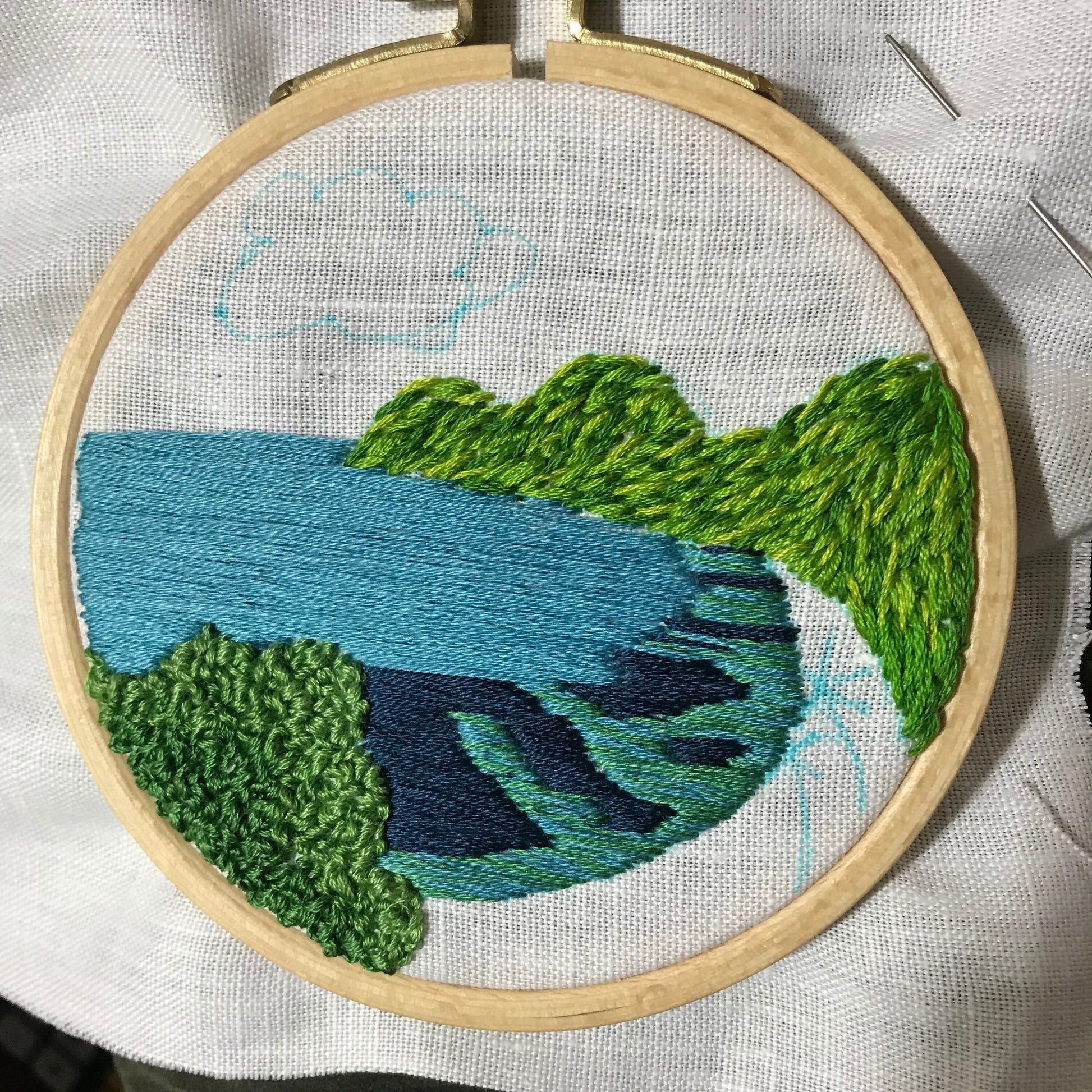 Hanauma Bay Embroidery design in progress.  Shows stitch detail of hills in green and water in blues blues and green.  All stitched on white linen with blue water soluble marker outlines for remaining details.