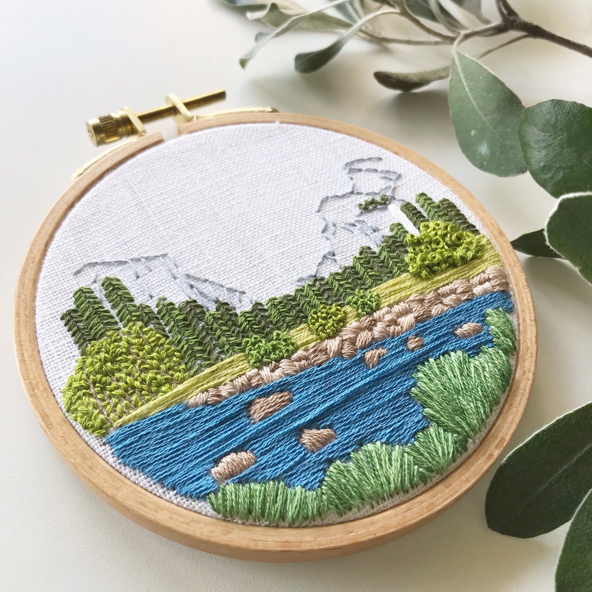 Completed Yosemite Valley Embroidery. 4 inch hoop has landscape scene on white linen.  Long blue stitches fill the river while shorter grey stitches make up the outline of the mountains.