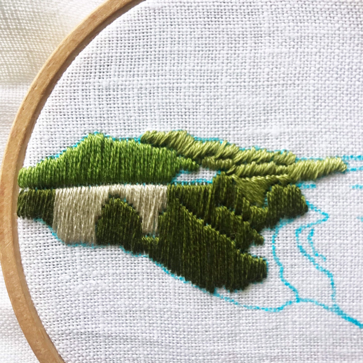 Big Sur Beginner Embroidery kit in progress, green hills and under color for bridge completed. Shows blue water soluble pen over white linen fabric.