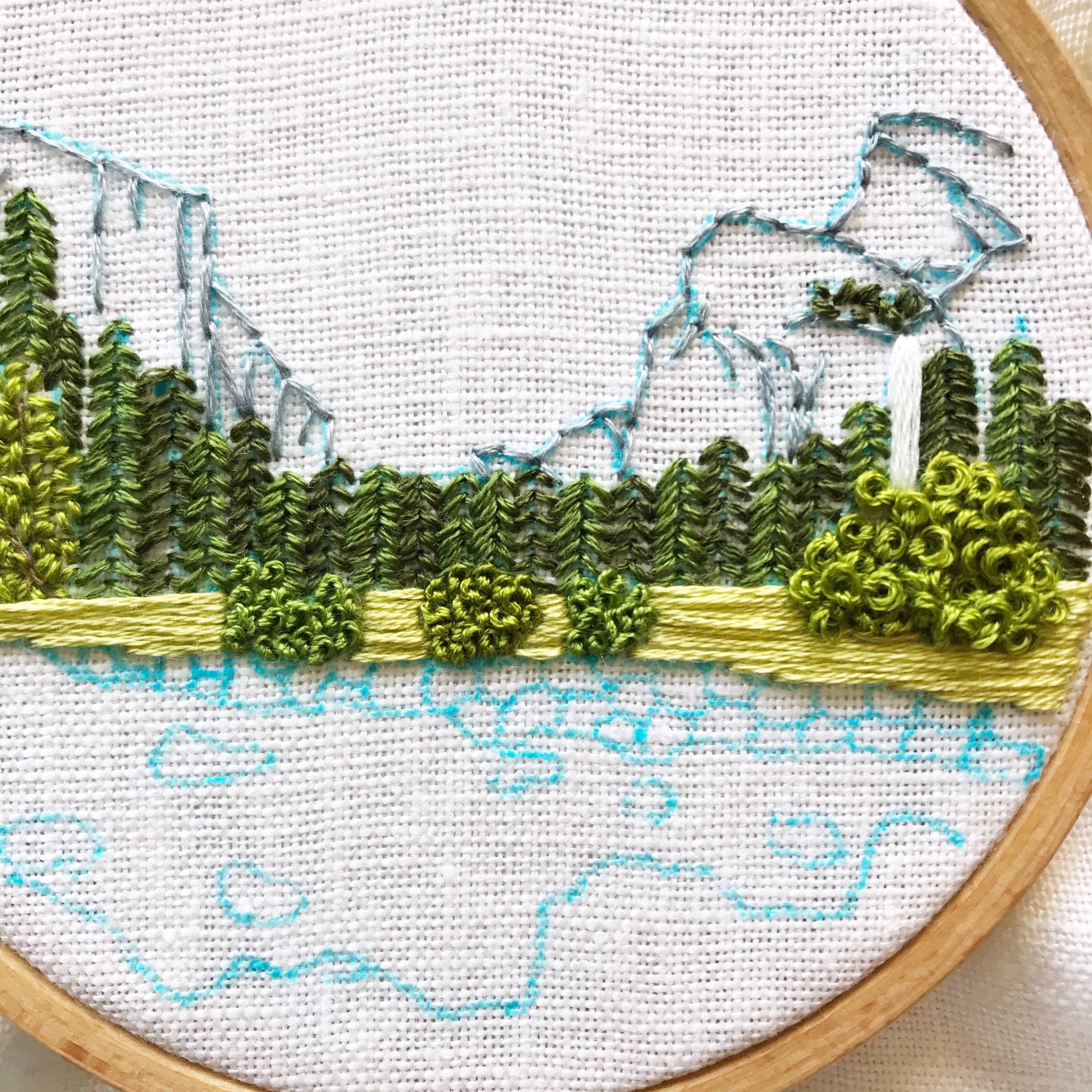 Yosemite Valley Embroidery Kit in progress. Mountains are stitched in grey over blue markings on white linen.  Various trees and shrubs are stitched in an array of greens.