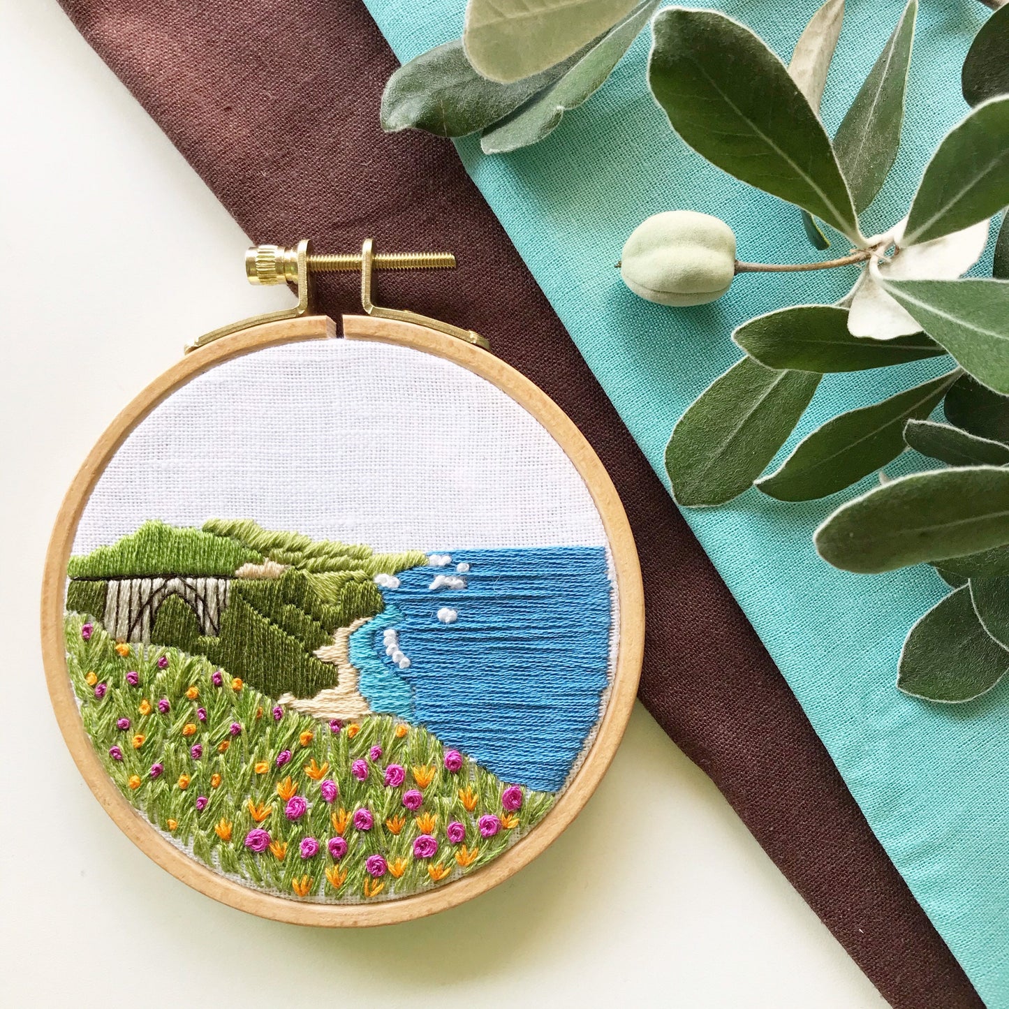 Big Sur California Hand Embroidery PDF. 13 page booklet gives easy to follow directions to complete this oceanside Modern Embroidery. Flowers in the foreground with HWY 101 Big Sur Bridge in the background. White waves crashing on beach.