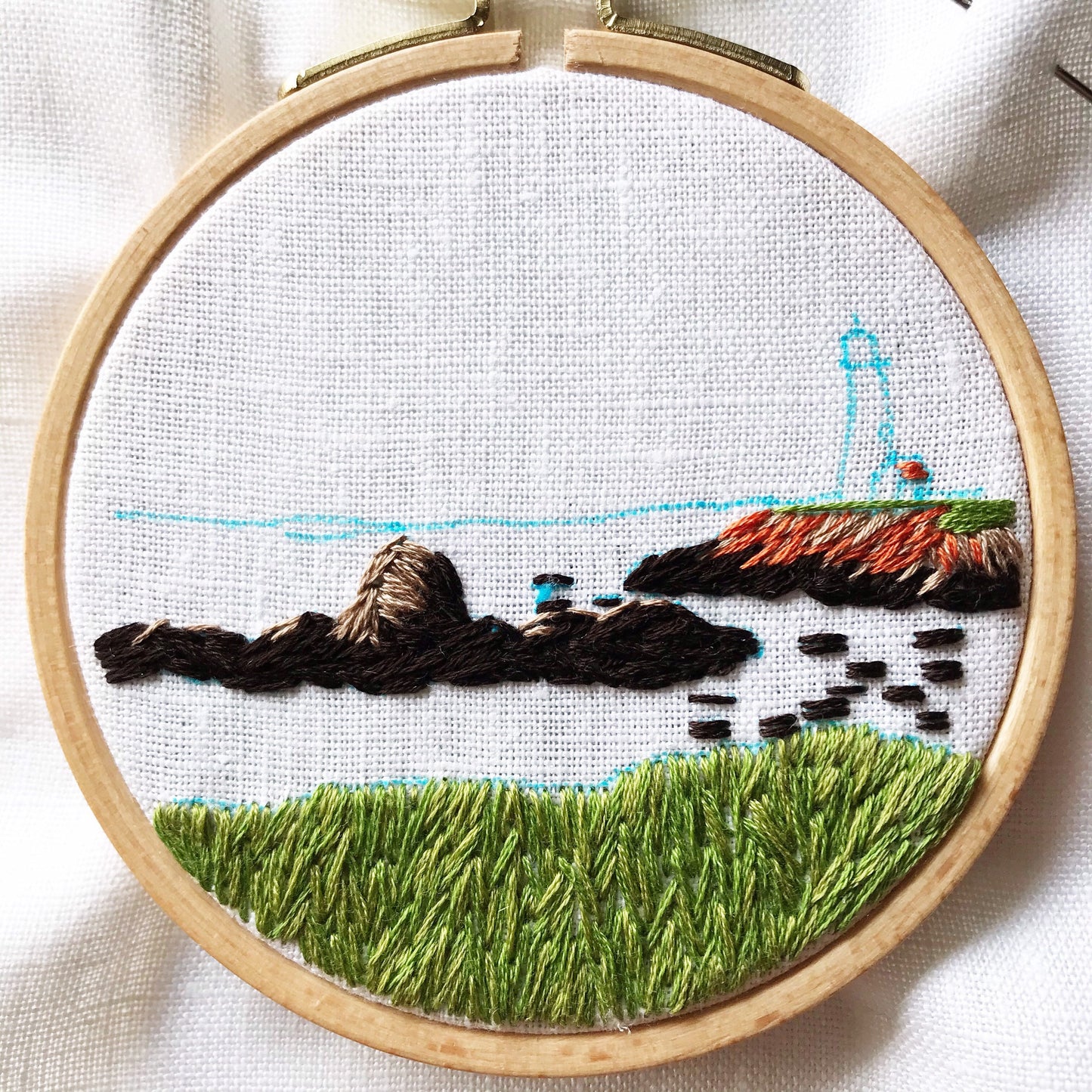 Lighthouse by the Bay Hand Embroidery in progress. Blue marker outlines stitch design. Grass and rocks have been completed in greens and browns.
