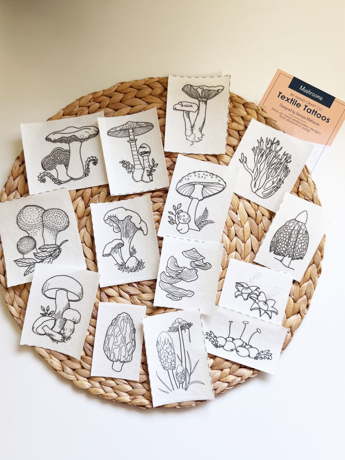 A set of small square fabric cards with black mushroom and botanical line drawings, arranged on a round woven placemat. A small card at the top right reads 'Textile Tattoos'.