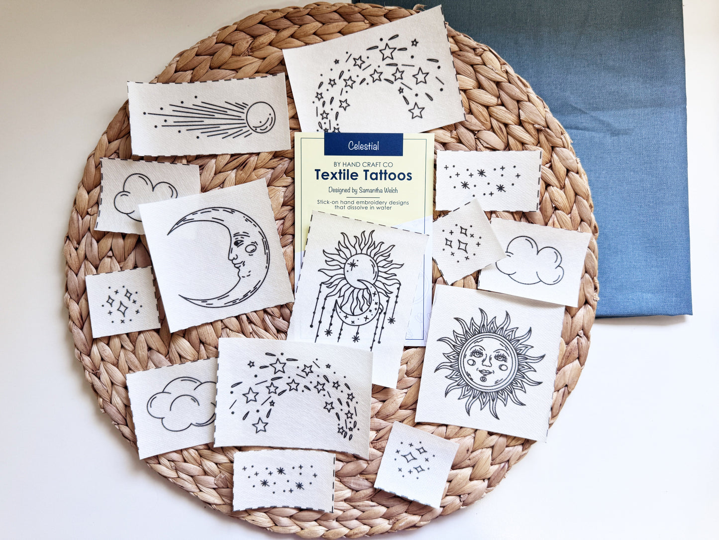 A flat-lay view of a woven straw tray holding multiple stick and stitch designs printed with black celestial designs like suns, moons, clouds, and stars. A yellow and blue paper card reading 'Textile Tattoos' is placed in the center, with a blue cloth underneath the tray.