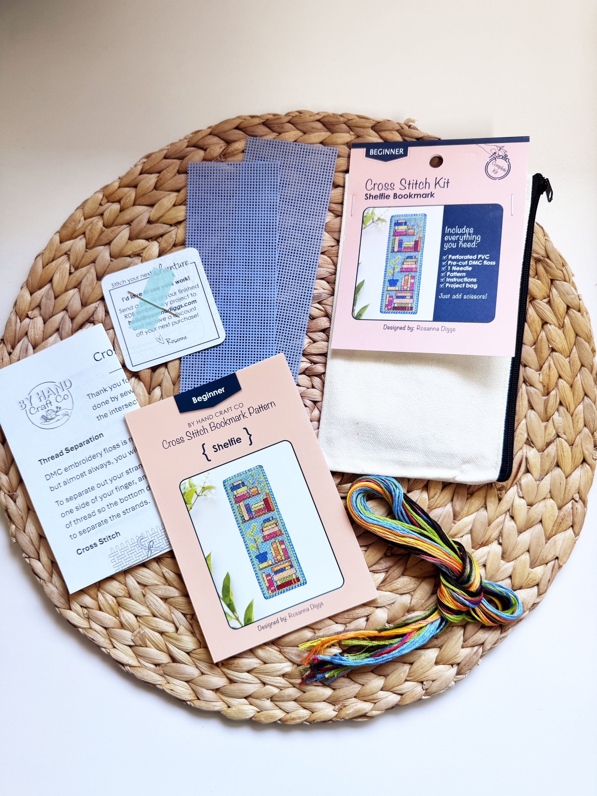 A cross stitch kit laid out on a woven round mat, including pattern cards, thread, fabric, a zip pouch, and a packaged cross stitch kit. 