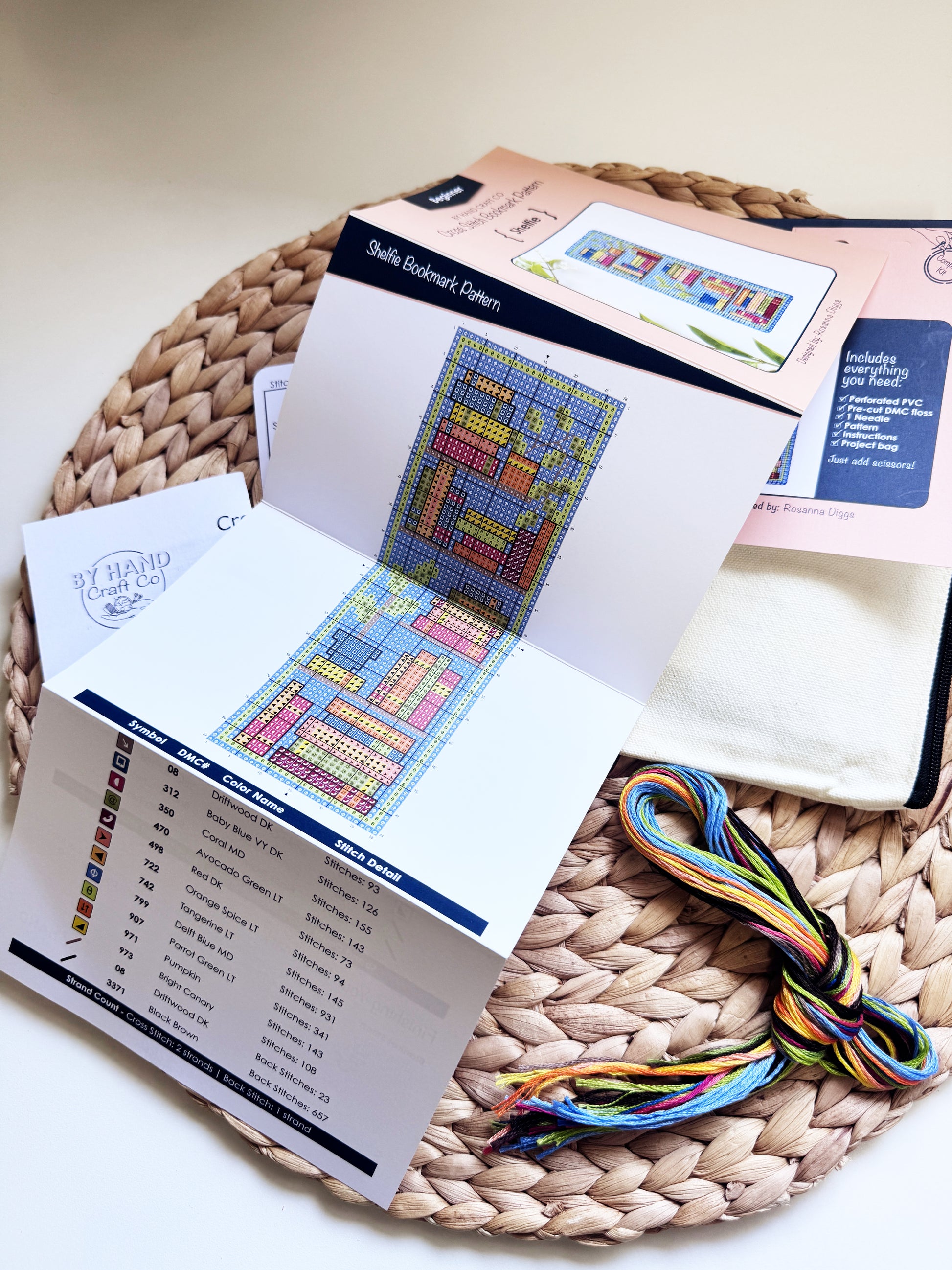 A cross-stitch kit laid out on a woven round mat, including an open pattern booklet with a colorful grid design, a list of thread colors, a folded white fabric piece, a pink instruction card, and a bundle of multicolored embroidery floss.