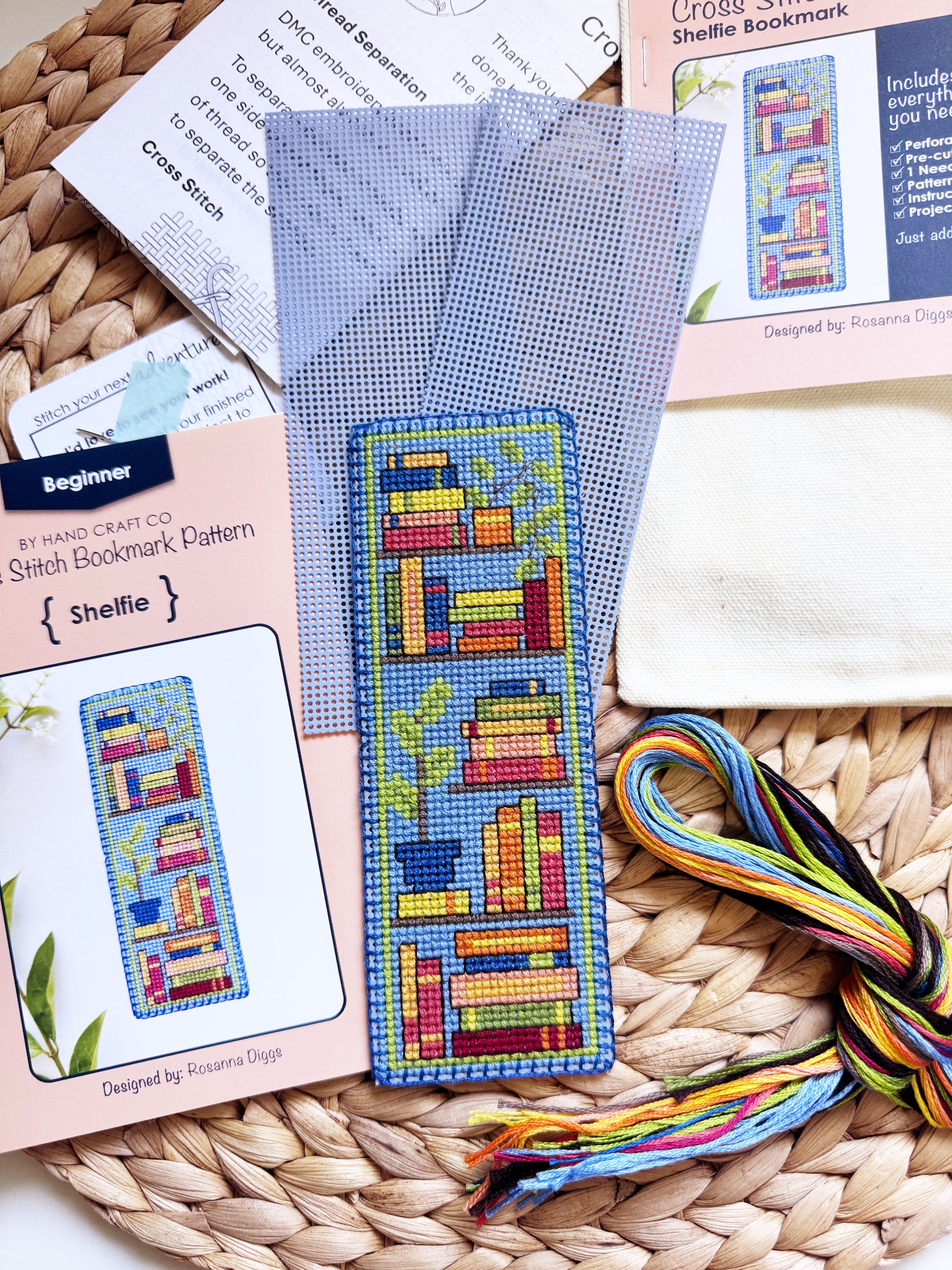 A cross-stitch bookmark kit laid out on a woven mat, including a finished sample bookmark, pattern cards, thread, and a white fabric square. The bookmark design features colorful books and a blue border.