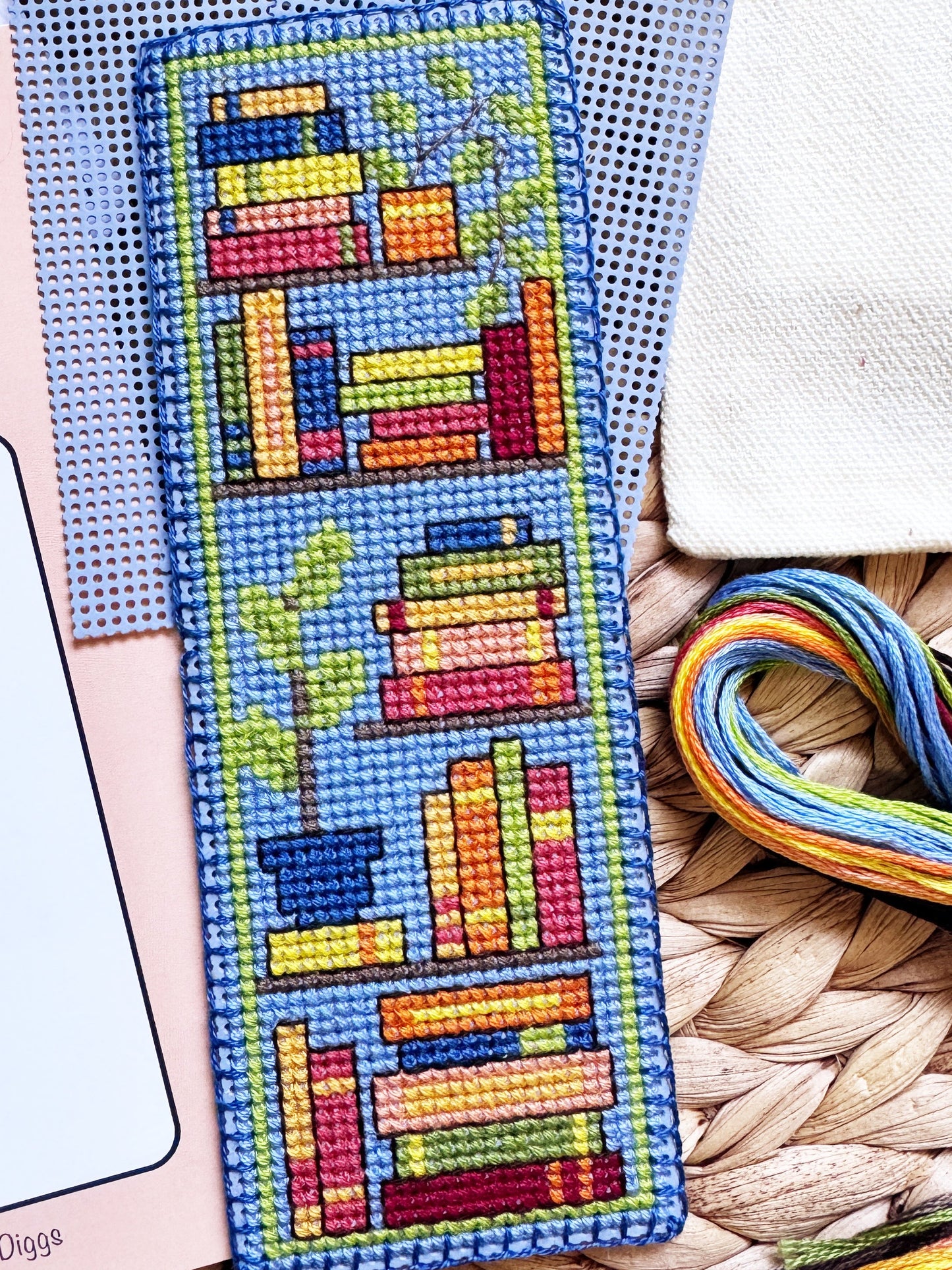 A completed cross-stitch bookmark featuring colorful book stacks and a potted plant on a blue background, displayed alongside its pink pattern booklet, blue Aida cloth, embroidery floss, and a woven mat.