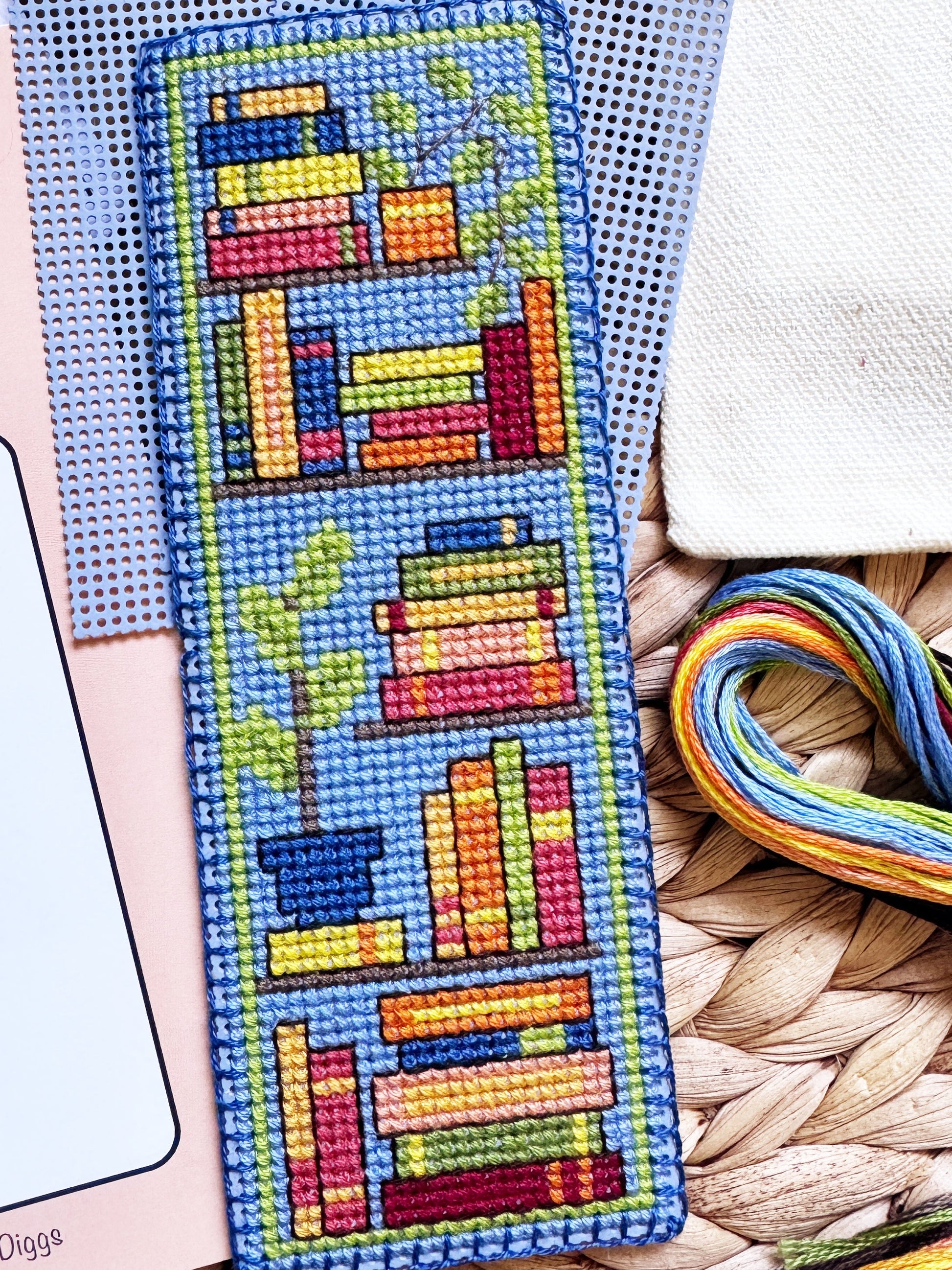 A completed cross-stitch bookmark featuring colorful book stacks and a potted plant on a blue background, displayed alongside its pink pattern booklet, blue Aida cloth, embroidery floss, and a woven mat.