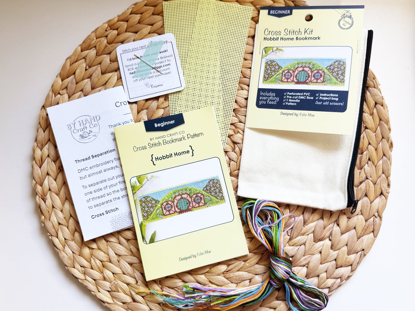 A cross stitch kit laid out on a round woven straw mat, including pattern cards, thread, fabric, and a finished sample piece. 