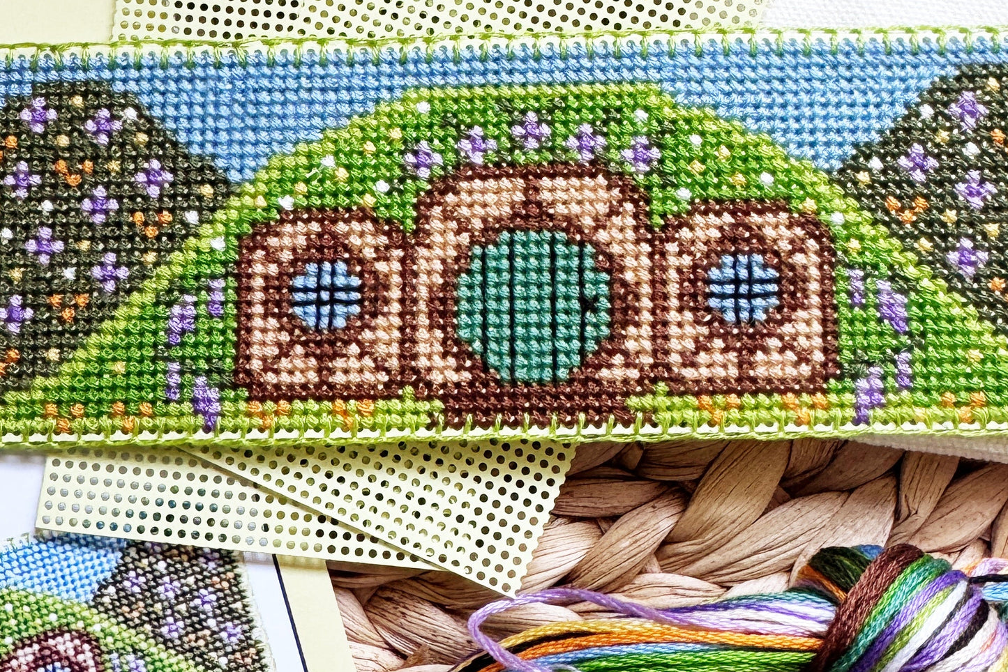 A Hobbit-themed cross-stitch kit laid out on a woven straw mat, showing the finished stitched design on a rectangular canvas, instruction sheets, thread skeins, and pattern cards.