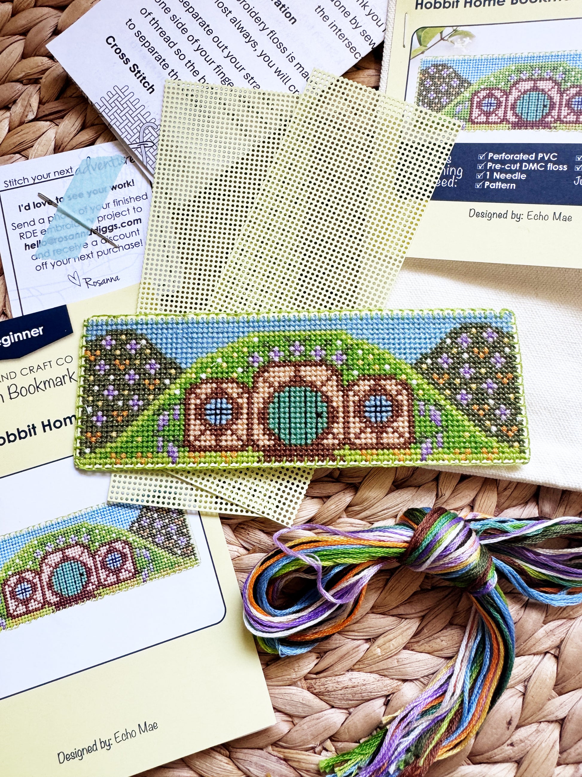 A Hobbit-themed cross-stitch kit laid out on a woven straw mat, showing the finished stitched design on a rectangular canvas, instruction sheets, thread skeins, and pattern cards.