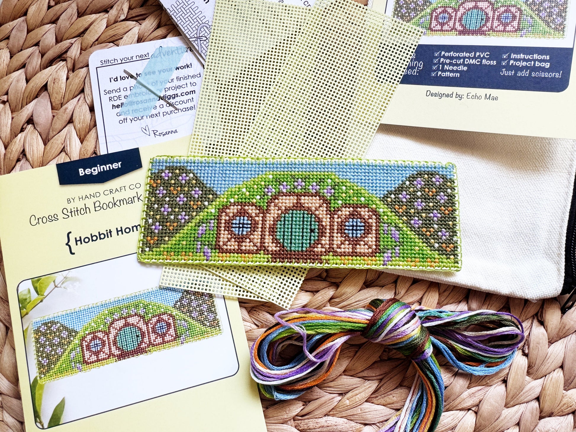 A cross-stitch kit laid out on a woven wicker surface, including a finished sample of a hobbit house, pattern cards, mesh fabric, a small pouch, and a bundle of multicolored thread.