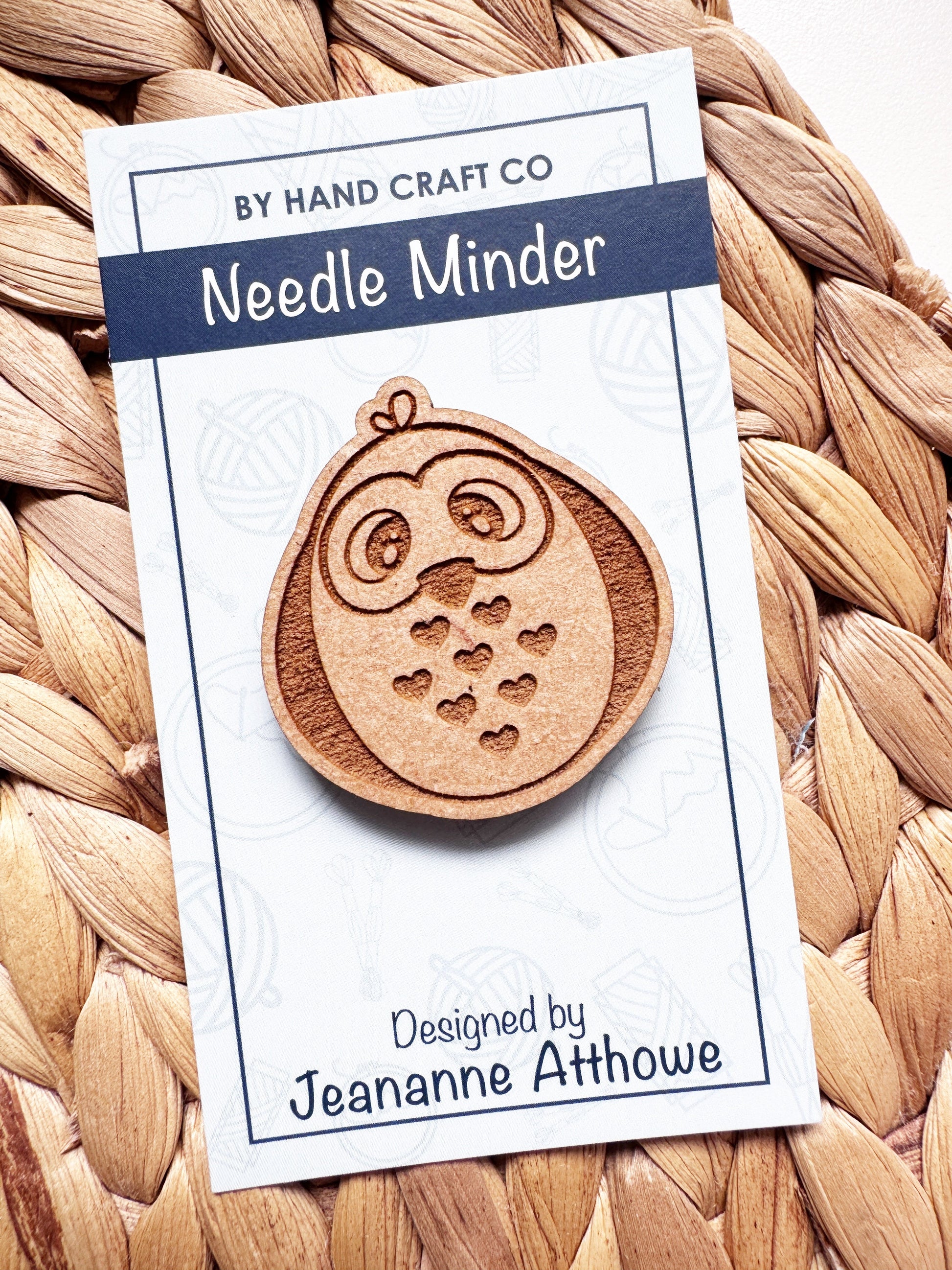 A wooden owl-shaped needle minder carded on a white backing card with blue text, placed on a woven straw mat. The card reads 'Needle Minder' and is designed by Jeananne Athowe.
