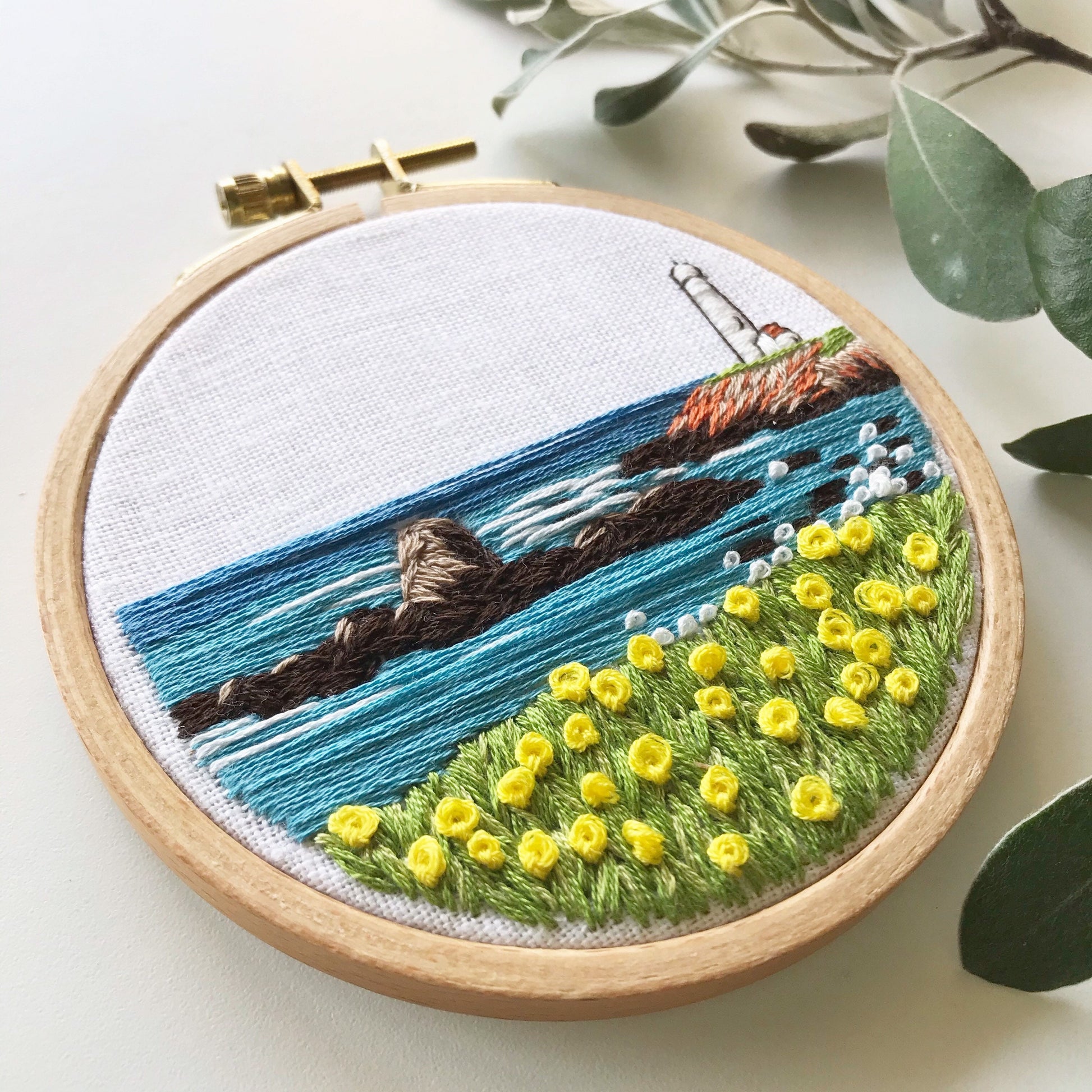 Lighthouse by the Bay PDF. Download includes easy to follow directions.  Hand Embroidery has lighthouse on the right hand side on cliff with rocks and ocean to the horizon.  Grass and flowers fill the foreground.